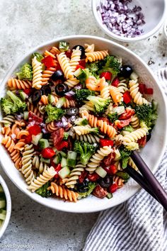 a white bowl filled with pasta salad next to two bowls full of vegetables and olives