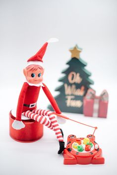 an elf sitting on top of a red cup next to a toy radio and christmas tree