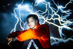 a man in an orange hoodie is holding his arms out while lightning strikes behind him