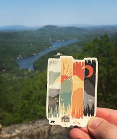 someone holding up a sticker with an image of mountains and trees in the background