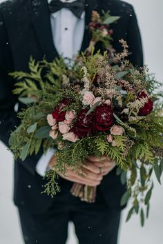 a man in a tuxedo holding a bouquet of flowers and greenery with snow falling on the ground