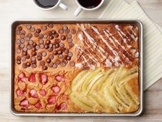 a pan filled with different types of food on top of a wooden table next to two cups of coffee