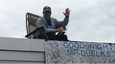 a man sitting on top of a roof waving to the crowd with his hand up