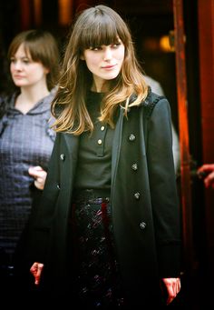 a woman with long hair wearing a black coat and dress standing in front of a door