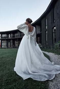 a woman in a white dress is standing on the grass near a building and looking off into the distance