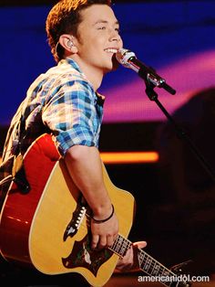 a young man holding a guitar and singing into a microphone while wearing a plaid shirt