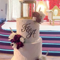 a three tiered wedding cake with red and white flowers on the side, sitting in front of a stage