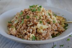 a white bowl filled with rice and vegetables on top of a wooden table next to a gold spoon