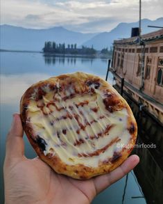 a hand holding a pizza with cheese and sauce on it in front of a body of water