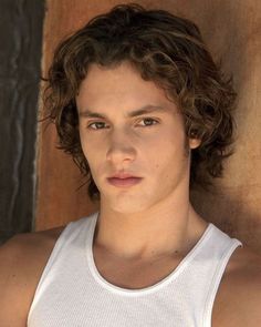 a young man with curly hair wearing a white tank top and looking at the camera