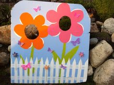 a painted wooden sign with flowers and butterflies on the front, sitting next to some rocks