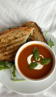 a white plate topped with a sandwich and bowl of tomato soup on top of a bed of lettuce