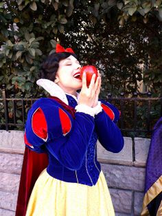 a woman dressed as snow holding an apple in front of her face while standing next to a stone wall