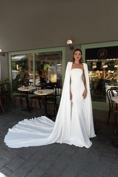 a woman standing in front of a restaurant wearing a white gown and cape with long train