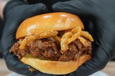 a hand in black gloves holding a sandwich with meat and onion rings on it's bun