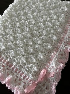 a white crocheted blanket with pink ribbon on the bottom and side, sitting on a black surface