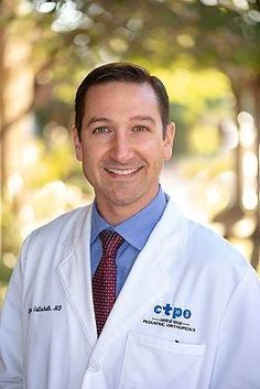 a man in a white lab coat and tie smiling at the camera with trees behind him