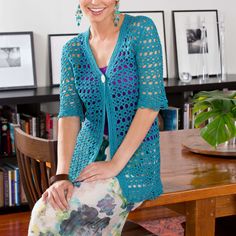 a woman sitting at a wooden table wearing a blue crocheted cardigan and smiling