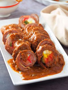 a white plate topped with meat rolls covered in sauce next to a bowl of vegetables