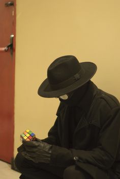 a man in a black hat and coat sitting on the floor with a rubix cube