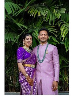 two people standing next to each other in front of some trees and palm leaves on the ground