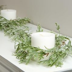 two white candles sitting on top of a table covered in green leaves and mossy branches