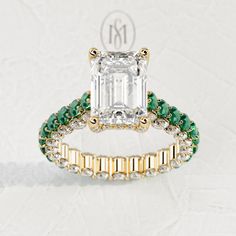 an emerald and diamond ring set on a white surface with the initials m in gold