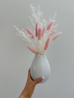 a hand holding a white vase with pink and white flowers in it on a gray background