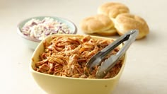 a yellow bowl filled with shredded meat next to rolls