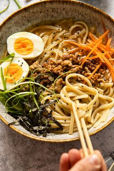 someone is holding chopsticks over a bowl of noodles with meat, egg and vegetables