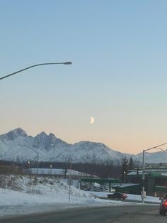 a car driving down the road in front of snow covered mountains and street lights at dusk