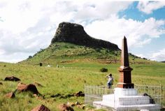 a man standing on top of a lush green field next to a tall rock formation