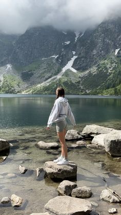 a woman standing on rocks in front of a lake with mountains in the back ground