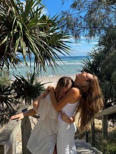 two young women hugging each other on the steps by the beach with palm trees in the background