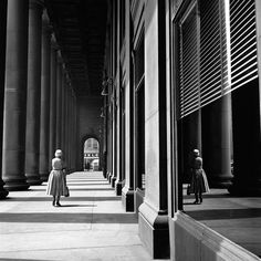 black and white photograph of woman walking down the sidewalk in an old building with columns