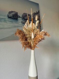 a white vase with dried plants in it on a table next to a wall and pictures