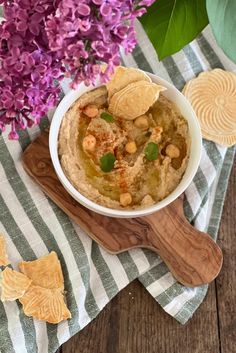 a bowl filled with hummus and chips on top of a wooden cutting board next to purple flowers