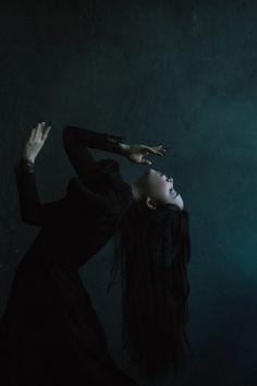 a woman with long hair standing in front of a dark background holding her hands up