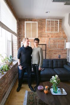 two people standing next to each other in front of a living room with brick walls