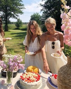 two women standing in front of a cake on a table with flowers and other desserts