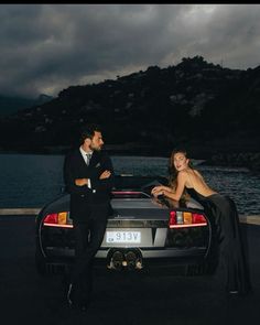 a man and woman leaning on the back of a sports car in front of a body of water