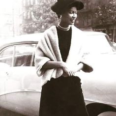 a black and white photo of a woman standing in front of a car wearing a hat