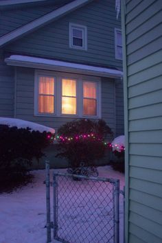 a house with christmas lights in the window and snow on the ground around it's fence