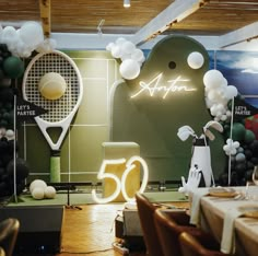 a room filled with balloons and tables covered in white table cloths next to a neon sign that reads 50