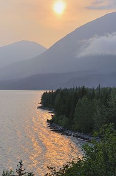 the sun is setting over some water and trees in front of a large mountain range