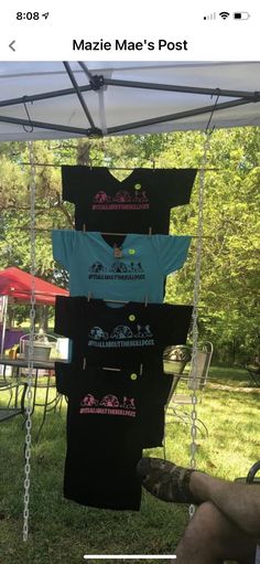 t - shirts hanging from a pole in the grass under a tent with people sitting around it