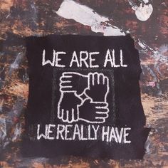 An unevenly cut, square, black fabric patch is on a paint-stained background. On the patch, there is an image of four hands in an interlocked square. Above the hands, there are the words, in all caps, "We Are All", beneath the hands in all caps are the words "We Really Have". Patch Design Ideas Punk, Punk Pins And Patches, Folk Punk Patches, Punk Patch Stencil, Punk Patches Diy Ideas, Scrap Punk, Punk Diy Ideas, Punk Patch Ideas, Punk Embroidery Ideas