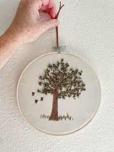 a hand is holding an embroidery hoop with a tree on it and bees flying around