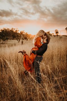 a woman in an orange jacket and cowboy hat is holding a baby while standing in tall grass