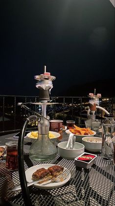 an outdoor table with food and drinks on it at night, overlooking the city lights